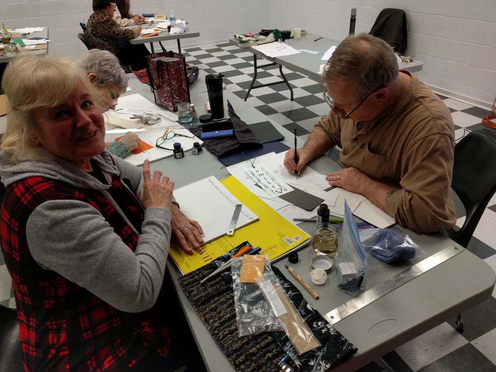 Instructor Rick Draffin demonstrates the use of an oblique holder to write Roundhand letters at an enthusiastic student's desk while taking a break from making weathergrams
